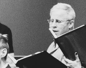 A black and white photo of E. Das Janssen singing during a concert with is face partially obscured by another choir member's music folder